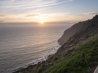 a person sitting on a hill overlooking the ocean and ocean waves at sunset time looking out over the ocean