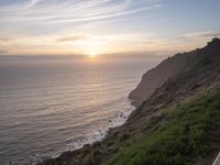 a person sitting on a hill overlooking the ocean and ocean waves at sunset time looking out over the ocean