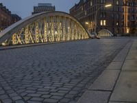 a bridge over a cobblestone street in a city at dusk with many buildings