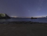 stars and a clear sky are seen over the ocean and mountains at night with rocks