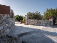 a stone fence sits near a stone building and gate in the country setting of a greek property