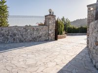 a stone fence sits near a stone building and gate in the country setting of a greek property