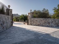 a stone fence sits near a stone building and gate in the country setting of a greek property