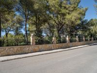 a long stone fence along the street in front of some trees and a bench near a fence