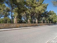 a long stone fence along the street in front of some trees and a bench near a fence