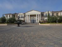 a stone plaza in front of a large mansion with a fountain in the middle surrounded by greenery