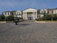 a stone plaza in front of a large mansion with a fountain in the middle surrounded by greenery