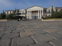 a stone plaza in front of a large mansion with a fountain in the middle surrounded by greenery