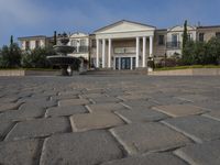 a stone plaza in front of a large mansion with a fountain in the middle surrounded by greenery