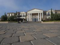 a stone plaza in front of a large mansion with a fountain in the middle surrounded by greenery