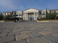 a stone plaza in front of a large mansion with a fountain in the middle surrounded by greenery