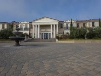 a stone plaza in front of a large mansion with a fountain in the middle surrounded by greenery