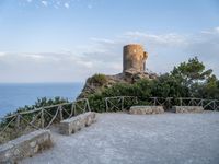 a stone wall and concrete walkway on the hillside near an old castle tower with a bench overlooking the sea