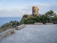 a stone wall and concrete walkway on the hillside near an old castle tower with a bench overlooking the sea