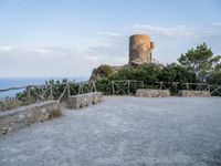 a stone wall and concrete walkway on the hillside near an old castle tower with a bench overlooking the sea