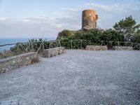 a stone wall and concrete walkway on the hillside near an old castle tower with a bench overlooking the sea