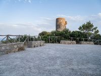 a stone wall and concrete walkway on the hillside near an old castle tower with a bench overlooking the sea