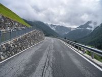 a road with a stone wall and metal railing, next to mountains and mountains are featured in this photo