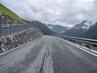 a road with a stone wall and metal railing, next to mountains and mountains are featured in this photo