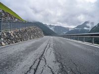 a road with a stone wall and metal railing, next to mountains and mountains are featured in this photo