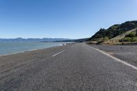 an empty road with a view of the ocean and mountains at high tide on a sunny day