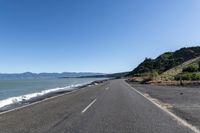 an empty road with a view of the ocean and mountains at high tide on a sunny day