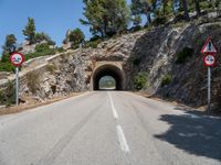 a tunnel leading into the mountains near some trees and road signs showing no left turn