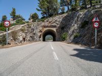 a tunnel leading into the mountains near some trees and road signs showing no left turn