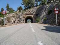 a tunnel leading into the mountains near some trees and road signs showing no left turn