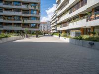 the paved walkway leads into an outdoor courtyard in urban city setting at daytime with people sitting on benches