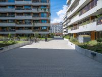 the paved walkway leads into an outdoor courtyard in urban city setting at daytime with people sitting on benches