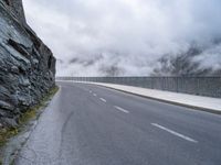 the road is straight and empty on this cloudy day by a mountain side wall with mist