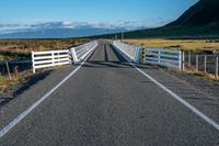 an empty road with a fence leading over the side of it and mountains in the distance