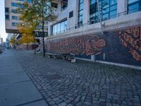 two park benches on a bricked sidewalk next to a building with a large art work on it