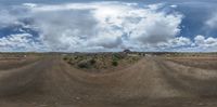 a wide panoramic picture of a dirt road in the desert with large clouds overhead