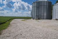 there is a large cement silo with several grain bins in the background that also has a sky filled with white clouds
