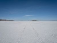 a single track in the middle of a vast expanse of snow with mountains and blue sky behind