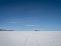 a single track in the middle of a vast expanse of snow with mountains and blue sky behind