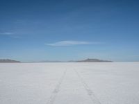 a single track in the middle of a vast expanse of snow with mountains and blue sky behind