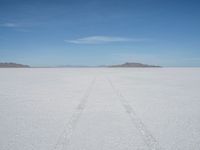 a single track in the middle of a vast expanse of snow with mountains and blue sky behind