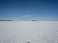 a single track in the middle of a vast expanse of snow with mountains and blue sky behind