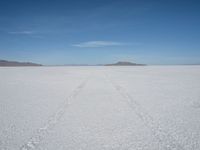 a single track in the middle of a vast expanse of snow with mountains and blue sky behind