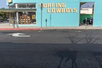 an empty road in front of a store that's on a corner with a blue wall and sign saying surfing cowboys