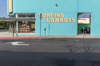 an empty road in front of a store that's on a corner with a blue wall and sign saying surfing cowboys