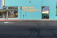 an empty road in front of a store that's on a corner with a blue wall and sign saying surfing cowboys