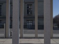 a street in front of a large building with concrete columns next to it and a sign attached to the wall reading the time