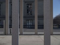 a street in front of a large building with concrete columns next to it and a sign attached to the wall reading the time