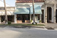 a small street lined with storefronts and palm trees in a corner area with blue, white, and tan awning