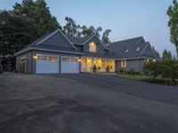 large suburban house with large driveway and garage with lit windows at dusk, next to trees