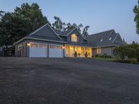 large suburban house with large driveway and garage with lit windows at dusk, next to trees